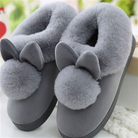 Chaussons d’intérieur gris doublés de fausse fourrure, avec pompons moelleux et petites oreilles décoratives, pour un confort chaud et cosy à la maison.