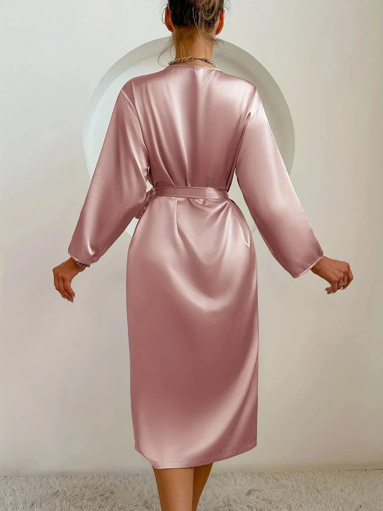 Vue de dos d’une femme portant un peignoir mi-long en satin rose poudré, à manches longues et ceinture nouée à la taille. Tissu fluide et brillant pour un style chic, confortable et élégant à la maison.