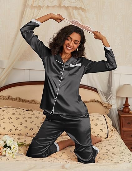 Femme joyeuse portant un pyjama en satin noir avec des détails en dentelle blanche, assise sur un lit. Elle sourit tout en tenant un bandeau de couleur rose au-dessus de sa tête, créant une atmosphère détendue et élégante. Le pyjama à manches longues et pantalon est à la fois chic et confortable, idéal pour se détendre à la maison.