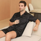 Homme assis sur un canapé portant le shorty en satin pour homme Sylvain, ensemble noir satiné à motifs subtils avec chemise à manches courtes et short assorti, tissu fluide et doux offrant un confort optimal, une allure élégante et une sensation agréable pour la détente à la maison ou la nuit.