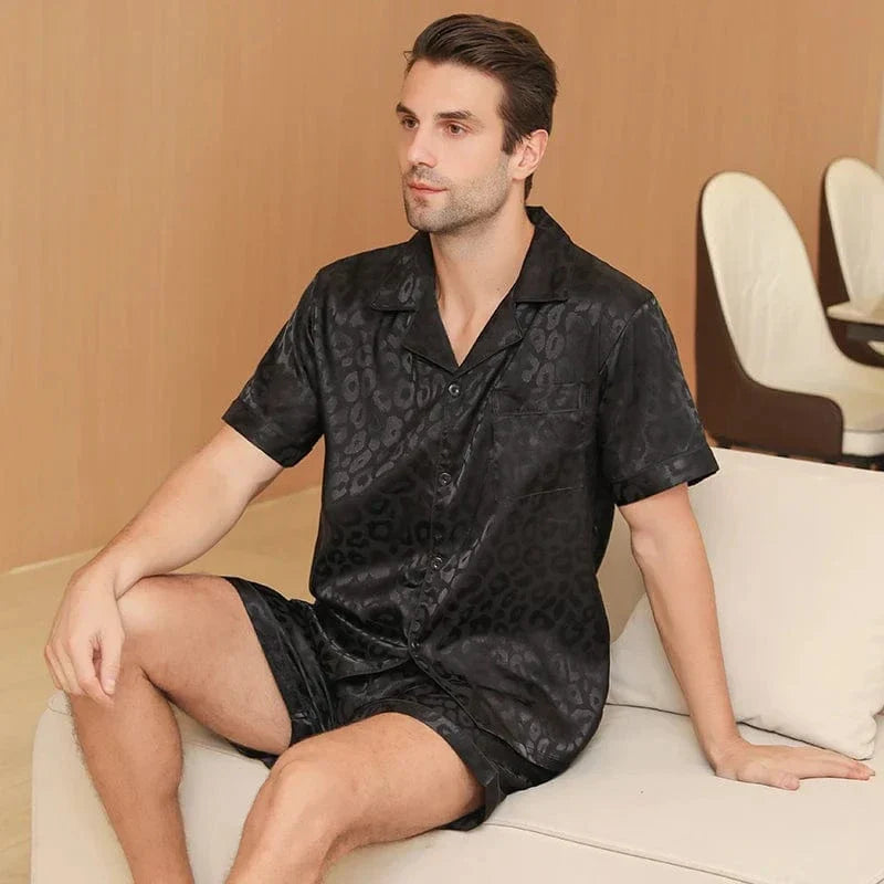 Homme assis sur un canapé portant le shorty en satin pour homme Sylvain, ensemble noir satiné à motifs discrets effet léopard, avec chemise à manches courtes boutonnée et short assorti, matière fluide et légère offrant un confort optimal et une allure élégante pour la maison ou la nuit.