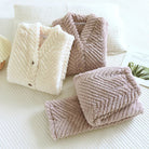 Deux ensembles de pyjamas pour femme en tissu pelucheux à motif chevron, un beige crème et un rose taupe, soigneusement pliés sur un lit blanc. Conçus pour offrir chaleur, douceur et confort absolu lors des soirées hivernales.