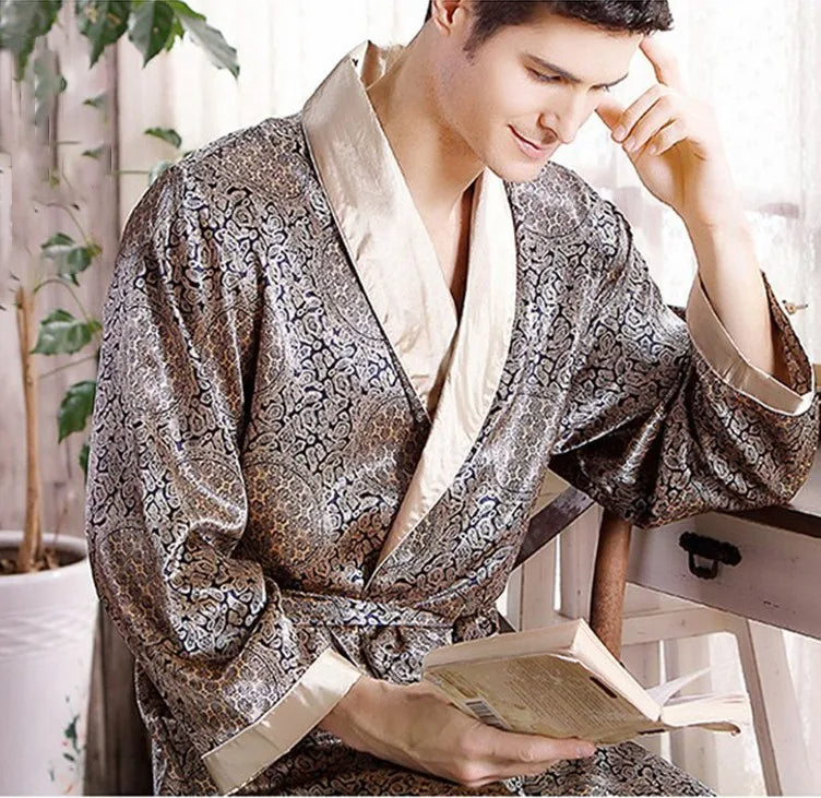 Homme portant le peignoir Tanguy, un modèle tendance et agréable en satin brillant à motifs dorés, assis près d’une fenêtre et lisant un livre dans une ambiance cosy.