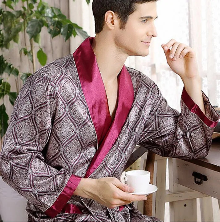 Homme portant le peignoir Tanguy, un modèle tendance et agréable en satin imprimé avec col et revers bordeaux, assis près d’une fenêtre lumineuse en tenant une tasse de café.
