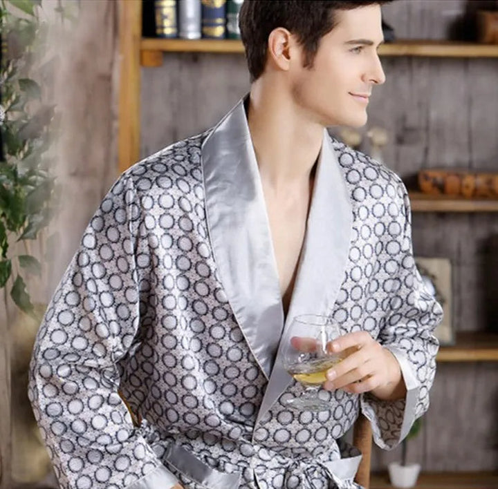 Homme portant le peignoir Tanguy, un modèle tendance et agréable en satin gris à motifs géométriques, assis dans un intérieur chaleureux tout en tenant un verre.