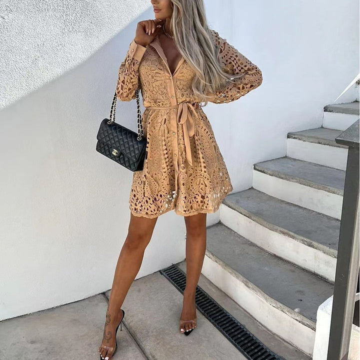 Une femme portant une robe en dentelle beige avec des manches longues et une ceinture satinée à la taille. Elle tient un sac à main noir de marque et pose élégamment devant un fond lumineux. Cette robe, raffinée et romantique, est parfaite pour des occasions spéciales, apportant une touche de sophistication à toute tenue.