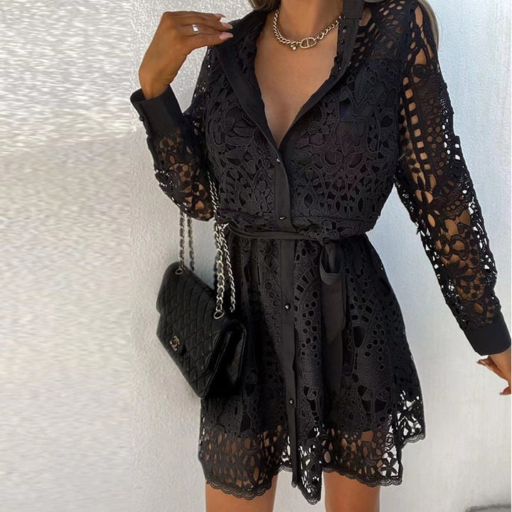 Une femme portant une robe en dentelle noire avec des manches longues, un col chemise et une ceinture satinée à la taille. Elle porte un sac à main noir de marque et un collier élégant, créant un look sophistiqué et raffiné. Cette robe est idéale pour des événements chics, offrant un mélange parfait d&