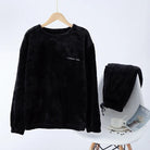 Pyjama femme Valérie composé d’un haut à manches longues en polaire noire suspendu sur cintre, accompagné d’un pantalon assorti posé sur une chaise blanche, ambiance cosy avec tasse, magazine et grains de café en arrière-plan. Ensemble chaud, doux et confortable pour l’hiver.