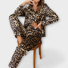 Femme assise sur une chaise portant un ensemble pyjama en satin imprimé léopard. Le haut à manches longues et le pantalon à coupe fluide sont parfaitement assortis, offrant un look chic et confortable. Elle pose avec une main sur son visage, mettant en avant l'élégance et le style du pyjama.