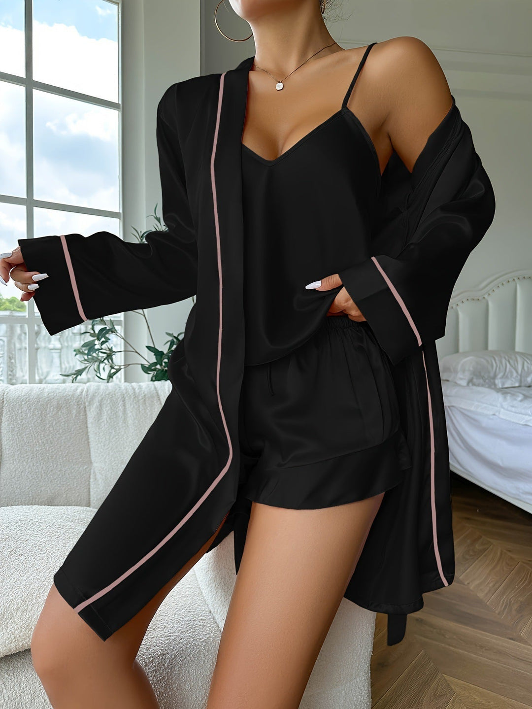 Pyjama femme Vivienne noir en satin, ensemble trois pièces doux et confortable avec caraco à fines bretelles, short fluide et peignoir long à manches longues, détails passepoilés rose poudré, porté dans une chambre.