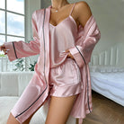 Pyjama femme Vivienne rose en satin, ensemble trois pièces doux et confortable avec caraco à fines bretelles, short fluide et peignoir long à manches longues, ceinture à nouer et finitions passepoilées noires, porté dans une chambre.
