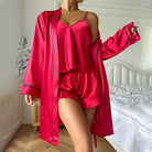 Pyjama femme Vivienne rouge en satin, ensemble trois pièces doux et confortable avec caraco à fines bretelles, short fluide et peignoir long à manches longues, ceinture à nouer et finitions passepoilées contrastantes, porté dans une chambre.
