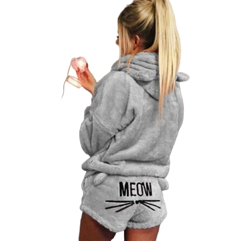 Femme portant le pyjama Whitaker, un ensemble en matière pelucheuse grise composé d’un haut à capuche avec oreilles de chat et d’un short assorti imprimé « MEOW » avec moustaches, offrant un style doux, chaud et amusant.