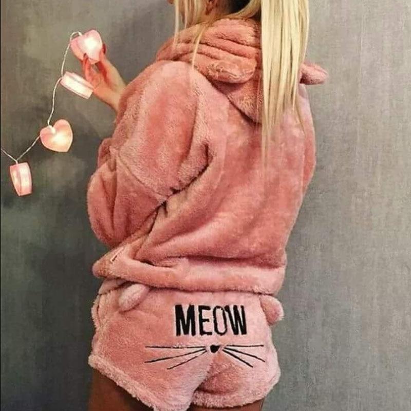 Femme portant le pyjama Whitaker, un ensemble en matière pelucheuse rose composé d’un haut à capuche avec petites oreilles de chat et d’un short assorti décoré du mot « MEOW » et de moustaches, offrant un style doux, chaud et ludique.