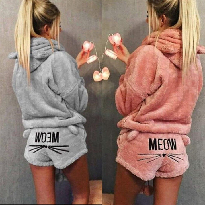 Deux versions du pyjama Whitaker pour femme, un ensemble doux et chaud en matière pelucheuse avec capuche à oreilles de chat et short assorti décoré du mot « MEOW » et de moustaches, présentées en gris et en rose pour un style à la fois confortable et amusant.
