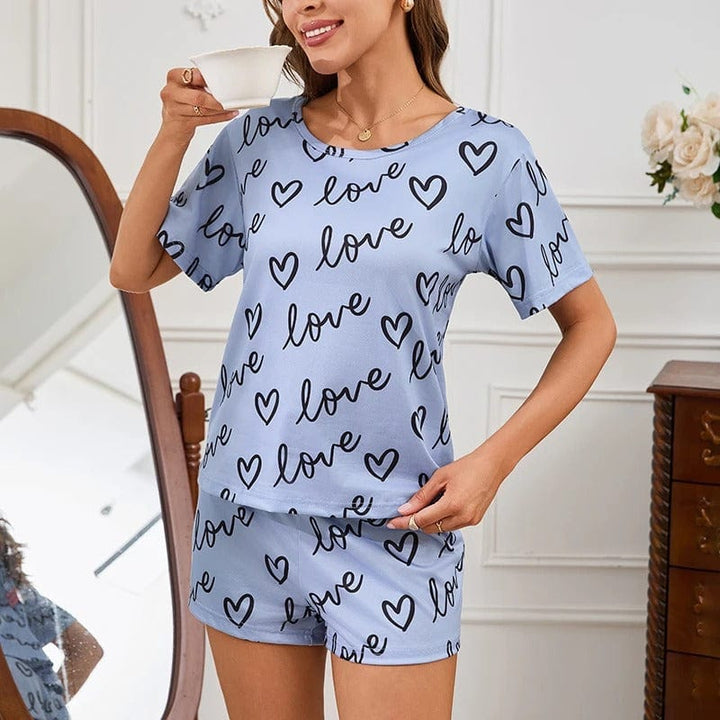 Femme souriante tenant une tasse, vêtue d’un pyjama court bleu clair à motifs “love” et cœurs noirs. Ensemble composé d’un t-shirt à manches courtes et d’un short assorti. Tenue douce, légère et agréable à porter, parfaite pour les nuits d’été ou les moments cocooning à la maison.