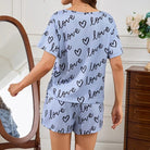 Vue arrière d’une femme portant un pyjama court bleu clair à imprimé "love" et cœurs noirs. Ensemble composé d’un t-shirt à manches courtes et d’un short assorti. Tenue confortable, douce et tendance, parfaite pour l’été ou les soirées détente à la maison.