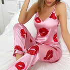 Pyjama satin femme Zéphyrine rose avec imprimé lèvres rouges, ensemble tendance et agréable avec caraco à fines bretelles et pantalon long satiné, porté assise sur un lit, tissu brillant et doux.