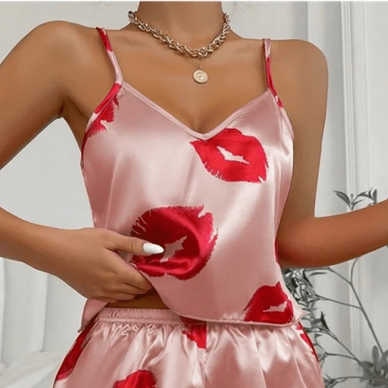 Gros plan sur un ensemble shorty en satin rose clair avec imprimé baisers rouges. Haut à fines bretelles avec encolure en V, porté avec un collier doré. Matière douce, coupe fluide et design tendance pour une tenue féminine et confortable, idéale pour les nuits estivales ou les moments de détente.